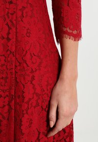 Punainen pitsimekko, jossa on kukkakuviot, kolme neljännesosaa hihat karvasaumoin ja tiukka siluetti. Käsi on asetettu kankaan päälle.