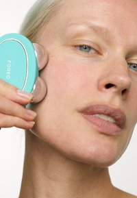Massager facial turquoise avec deux applicateurs en silicone arrondis et lisses, tenu contre la joue. La peau est claire, montrant des textures subtiles.