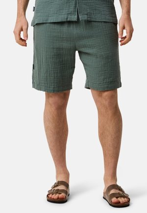 Shorts verts texturés avec une taille élastique, des poches latérales et une coupe décontractée, associés à des sandales marron à deux lanières sur des pieds nus.