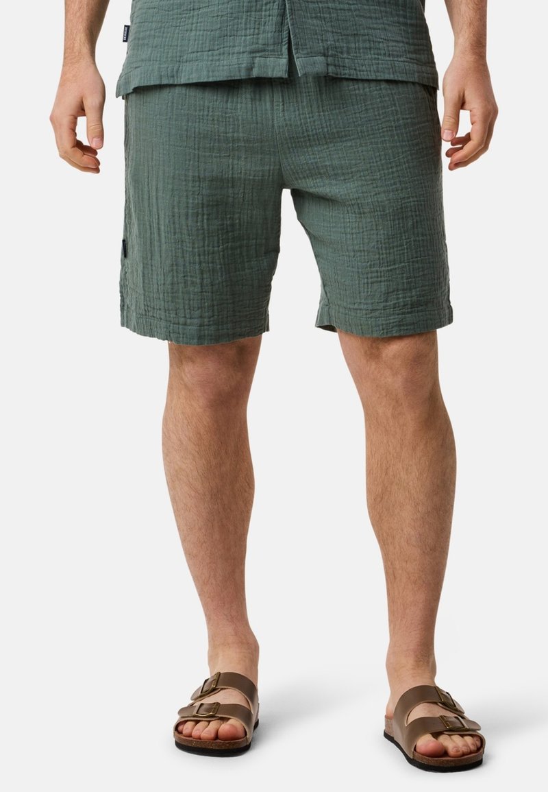Shorts verts texturés avec une taille élastique, des poches latérales et une coupe décontractée, associés à des sandales marron à deux lanières sur des pieds nus.