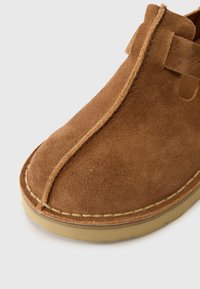 Brun mockasin i suède med rund tå, synlig sömnad och gummisula. Designen har en sidokut för enkel påtagning.