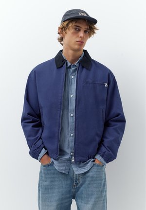 Ung mand iført en marineblå jakke med sort krave, stribet blå skjorte med knapper, denimjeans og sort kasket, stående med hænderne i lommerne.