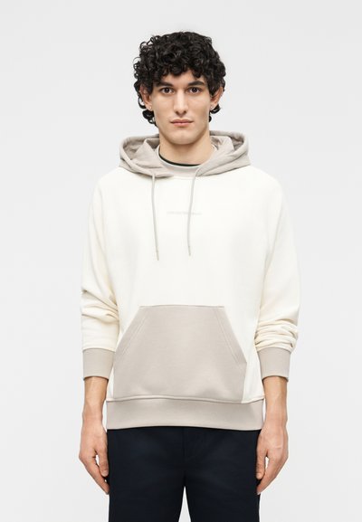 Νέος άνδρας με σκούρα σγουρά μαλλιά, φορώντας μπεζ και λευκό φούτερ με κουκούλα και μπροστινή τσέπη, στέκεται μπροστά σε απλό φόντο.