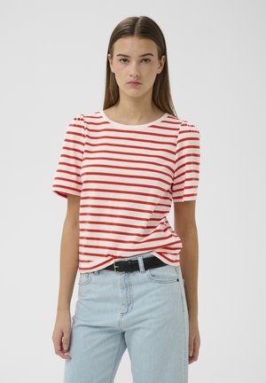 Vrouw met recht bruin haar die een wit en rood gestreept topje met korte mouwen draagt en lichtblauwe spijkerbroek met een zwarte riem.