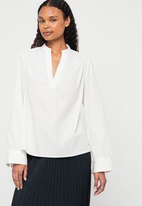 Bluză albă cu decolteu în V, mâneci lungi și încheieturi manșonate. Construită dintr-un material neted. Asortată cu o fustă neagră plisată.