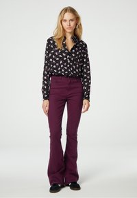 Zwarte bloemen overhemdbloes met witte en paarse bloemen, gecombineerd met hooggerimpelde pruimenkleurige flared pantalons en zwarte suède schoenen.