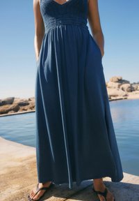 Robe maxi bleu foncé. Caractéristiques : corsage en maille, jupe fluide et poches latérales. Portée avec des tongs noires devant un décor d'eau rocheuse.