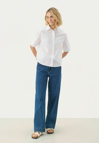 Witte overhemdd met korte mouwen, gecombineerd met hoge, wijde blauwe jeans en beige sandalen met een decoratief accent.
