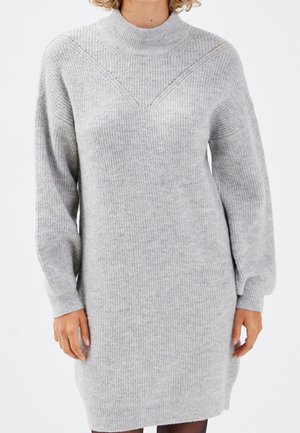 Femme portant une robe-pull à manches longues en maille côtelée gris clair, avec un col montant et un motif chevron subtil près des épaules.