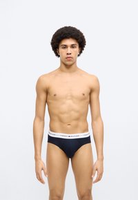 Μαύρο ανδρικό εσώρουχο με λευκή ζώνη μέσης, με το λογότυπο "TOMMY HILFIGER". Το μοντέλο στέκεται όρθιο, παρουσιάζοντας ουδέτερη μπροστινή όψη.