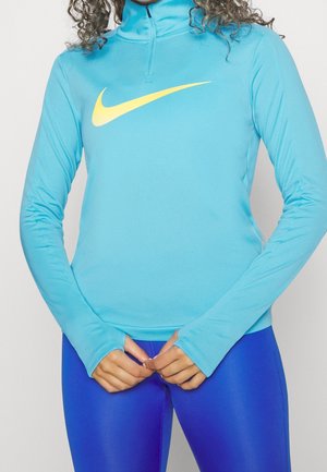 Osoba nosząca jasnoniebieski top z długim rękawem zapinany na zamek z żółtym logo Nike oraz błyszczące królewsko niebieskie legginsy, ręce splecione na wysokości talii.