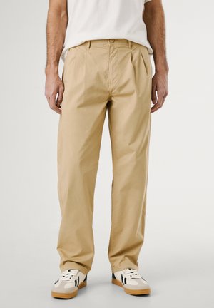 Pantaloni a pieghe khaki realizzati in tessuto leggero, con vestibilità dritta e tasche laterali, abbinati a sneaker bianche e nere.