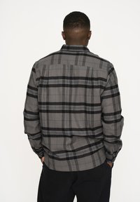 Camicia di flanella a scacchi grigia con motivi neri e blu, dettagli ai polsini con bottoni e orlo dritto. Il materiale sembra morbido e texture.