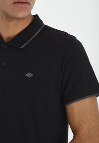 Černé polo tričko s límečkem, třemi knoflíky a šedými akcenty na límečku a rukávech. Obsahuje malé vyšívané logo.