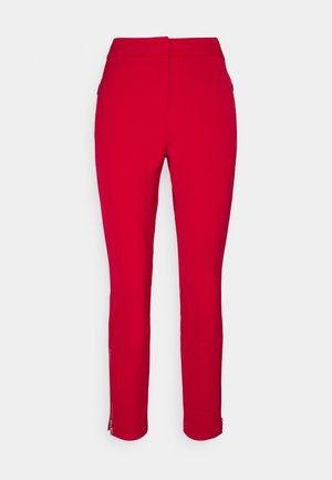Czerwone spodnie typu slim fit z zamkami po bokach i przy kostkach, pokazane od tyłu na jednolitym jasnym tle.