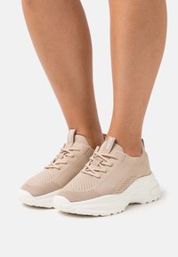 Beige stickade sneakers med en strukturerad ovandel, vit sula och snörningsdesign. Har en mjuk krage och ett varumärkesmärke på tungan.