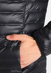 Veste matelassée noire avec une texture lisse et brillante, présentant des coutures horizontales et une main dans la poche latérale. Les poignets élastiques sont visibles.