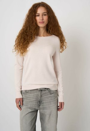 Vrouw met lang, krullend haar, gekleed in een lichtbeige trui en vervaagde grijze spijkerbroek, staand tegen een effen lichte achtergrond.