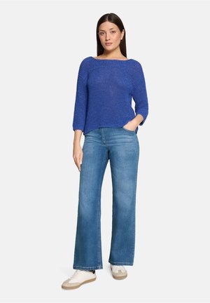 Frau steht mit einer Hand in der Tasche, trägt einen langärmeligen blauen Strickpullover, weite blaue Jeans und weiße Turnschuhe vor weißem Hintergrund.