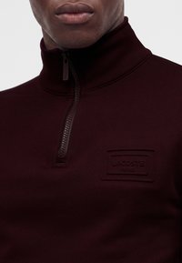 Homme portant un pull zippé bordeaux foncé avec un col montant et un discret écusson du logo Lacoste sur la poitrine.