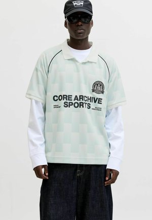 Homme portant une casquette noire, des lunettes de soleil foncées, une chemise blanche à manches longues sous un polo de sport vert clair avec du texte noir, et un jean large foncé.