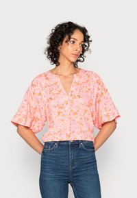 Blusa floral rosa com mangas curtas e largas, decote em V e detalhes de botões na barra. O tecido apresenta pequenos padrões de flores em laranja e branco.