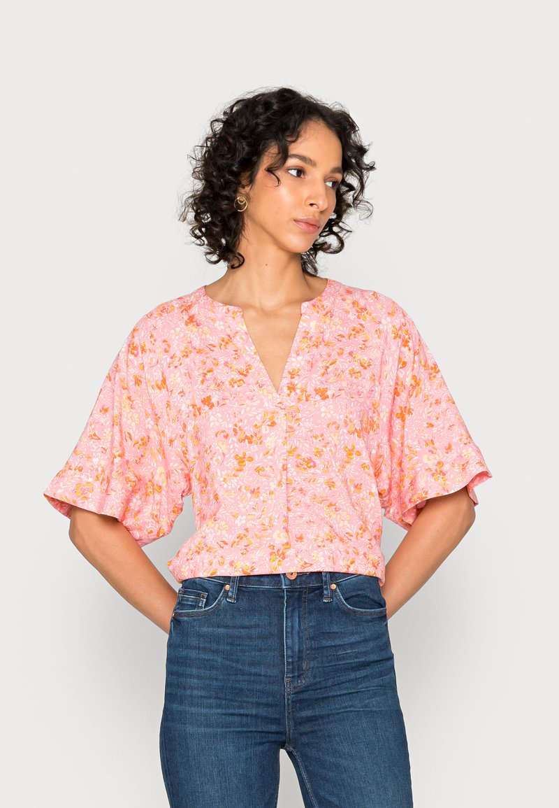 Blusa floral rosa com mangas curtas e largas, decote em V e detalhes de botões na barra. O tecido apresenta pequenos padrões de flores em laranja e branco.