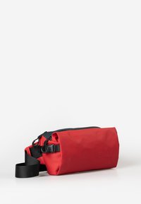 Compacte rode en zwarte schoudertas met een verstelbare zwarte strap en ritssluiting, rustend op een witte ondergrond.