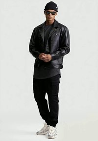 Uomo che indossa un berretto nero, occhiali da sole, giacca di pelle nera, pantaloni cargo neri, maglietta grigia e sneakers bianche, in piedi contro uno sfondo bianco.