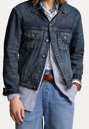 Persona vistiendo una chaqueta de mezclilla azul oscuro sobre una camisa a rayas azul y blanca, pantalones azul claro, cinturón de cuero marrón y reloj de pulsera con correa marrón.