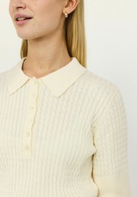 Suéter tipo polo color crema con patrón de tejido texturizado, tapeta de botones y cuello suave. Presenta un diseño entallado y detalles acanalados en las mangas.