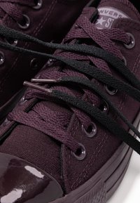 Lila canvas sneakers med glansigt tåhuvud, svarta snören och cirkulära öglor. Har texturerad tyg och kontrasterande detaljer.