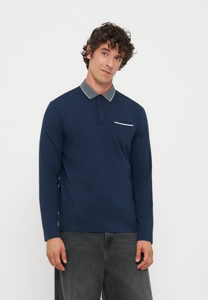 Homme aux cheveux bouclés portant un polo bleu marine à manches longues avec un col à motif et un pantalon gris foncé, debout devant un fond uni.