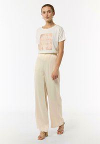 Kremowa koszulka z teksturowanym nadrukiem "WYZWÓL SWÓJ UMYSŁ", w zestawieniu z beżowymi spodniami z szerokimi nogawkami. Modelka nosi jasne sandały na słupku.