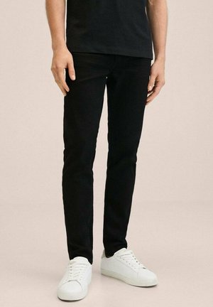 Personne portant un pantalon noir slim et des baskets blanches basses, debout devant un fond clair uni.