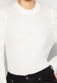 Pull en maille blanche côtelée avec col rond et manches longues bouffantes. La texture du tissu est douce et extensible. Associé à un jean noir taille haute.