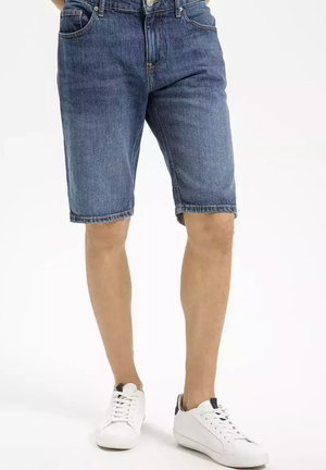 Szorty dżinsowe w średnim niebieskim kolorze o klasycznym kroju, z pięcioma kieszeniami i postrzępionym dołem, zestawione z białymi sneakersami.