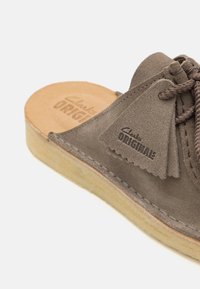 Gray suede sandal with a rounded toe, leather sole, and decorative tassel detail. Logo embossed on the insole. Visible stitching accents.