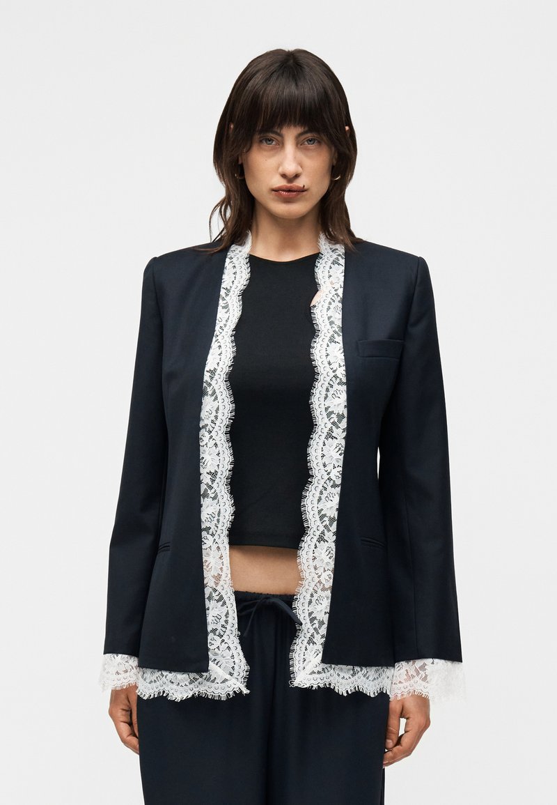 Γυναίκα με σκούρα μαλλιά μέχρι τους ώμους φοράει μαύρο σακάκι και παντελόνι με λευκή δαντέλα πάνω από μαύρο crop top, στέκεται μπροστά σε απλό φόντο.