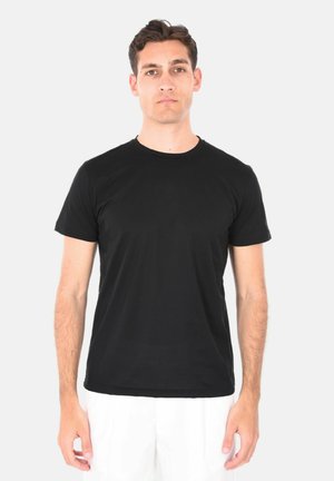 Uomo con capelli corti e scuri che indossa una t-shirt nera a maniche corte semplice e pantaloni bianchi, in piedi davanti a uno sfondo chiaro.