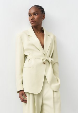 Γυναίκα που φοράει ανοιχτό μπεζ σακάκι με ζώνη και παντελόνι με φαρδιά μπατζάκια, στέκεται μπροστά σε λευκό απλό φόντο, κοιτώντας προς το πλάι.