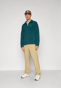 Hombre con gorra marrón, chaqueta con cremallera de color verde azulado, pantalones beige y zapatillas blancas, de pie contra un fondo claro sencillo.
