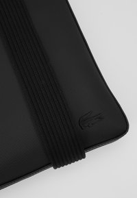 Housse d’ordinateur portable noire texturée avec large bande élastique et logo crocodile embossé dans le coin inférieur sur fond blanc.