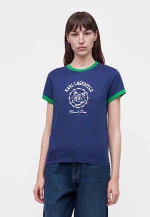 Kvinde med glat brunt hår iført en blå T-shirt med grøn kant og en hvid kattegrafik samt tekst foran, kombineret med blå jeans.