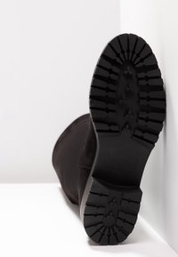 Bota negra con un upper de tela texturizada y una suela de goma negra resistente que presenta profundos surcos para tracción. Suela vista desde abajo.