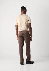 Beige kortermet skjorte kombinert med brune bukser med elastisk midje, med sidelommer og baklomme-detaljer, iført svarte sko.