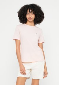 Camiseta de algodón rosa claro con mangas cortas, cuello redondo y pequeño detalle de logo en el pecho, combinada con pantalones cortos blancos.