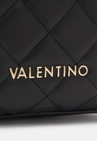 Mustan tikatun nahan päällä on kultainen "VALENTINO"-logo. Siinä on timantinmuotoinen kuvio ja sileä pinta.