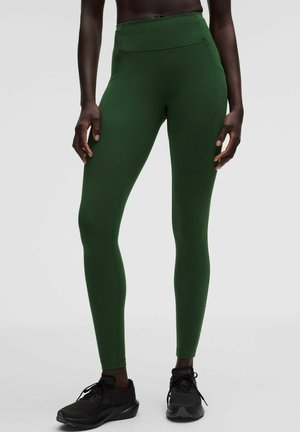 Osoba nosząca ciemnozielone legginsy z wysokim stanem i czarne buty do biegania, stojąca na jednolitym szarym tle.