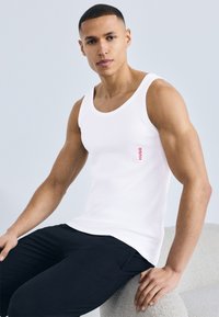 Νεαρός άνδρας με λευκό αμάνικο τοπ με κόκκινο κείμενο «HUGO», καθισμένος σε ανοιχτόχρωμη υφασμάτινη καρέκλα, φοράει μαύρο παντελόνι.